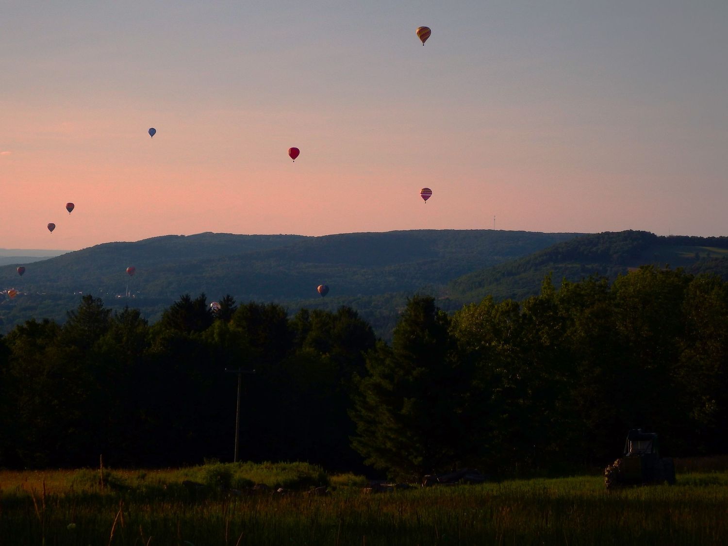 Hot air balloons float over a mountain landscape at sunset with silhouetted trees in the foreground.
