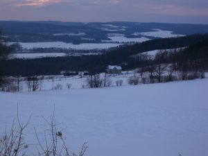 Snow-covered rolling hills and forests stretch across a winter landscape at dusk.