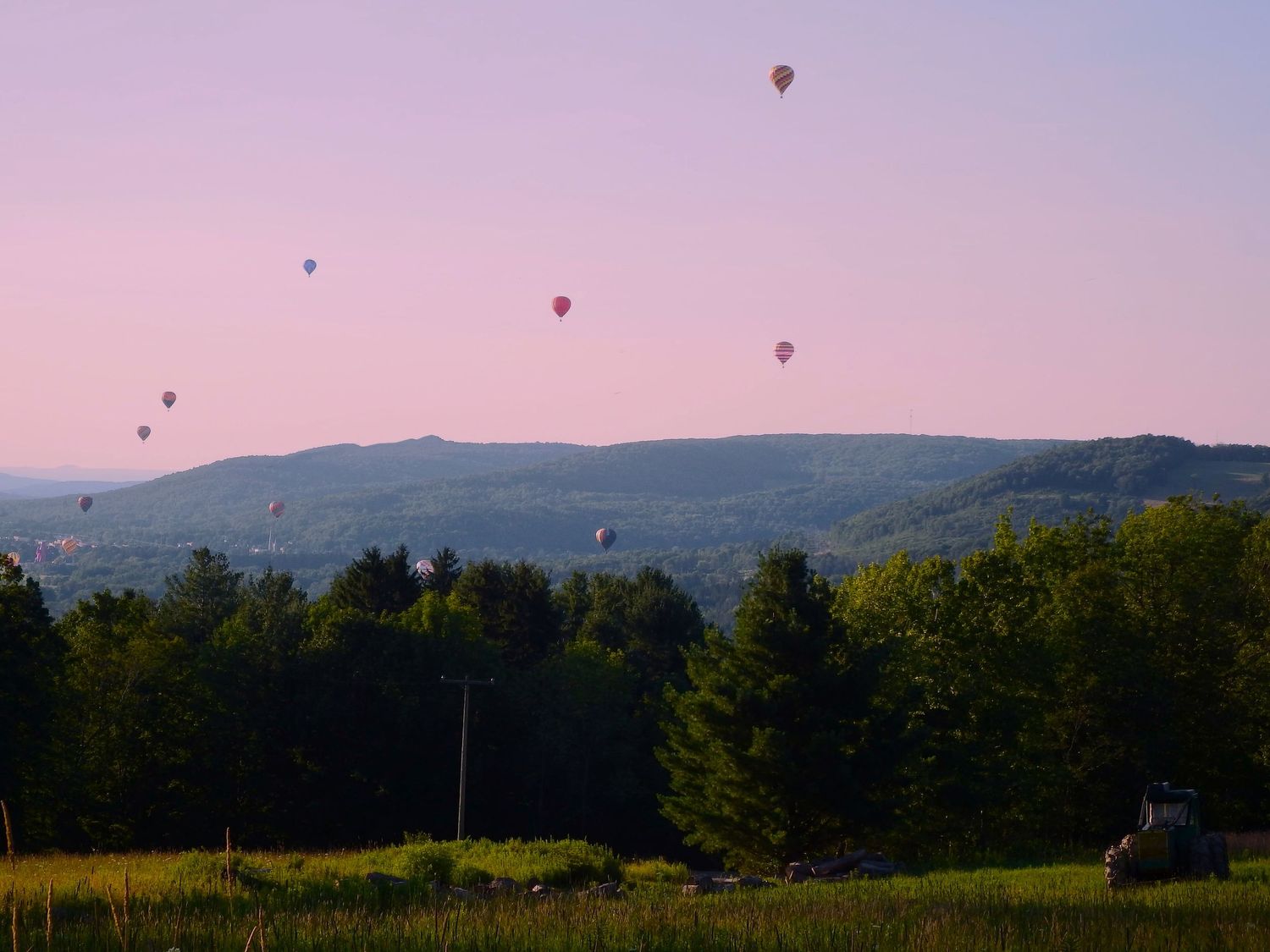Colorful hot air balloons drift across a pink sky above rolling hills and forests at dusk.