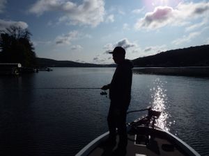 Silhouetted figure fishing from boat on calm lake during sunset.