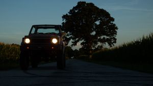 A vehicle with headlights on silhouetted against a dusky sky with a large tree in the background on a rural road.