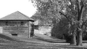 Historic wooden blockhouse structure with sloped roof sits among autumn trees in black and white.
