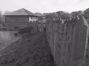 Black and white image of a wooden blockhouse with tall wooden stockade fence extending from its side.