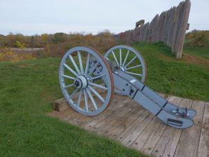 A light blue cannon sits on a wooden platform against a backdrop of wooden fortification walls.