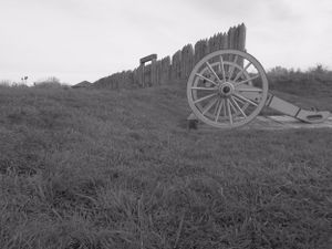 Black and white photo of a historic cannon positioned near wooden fortification walls on a grassy hill.