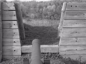 View through a gun port in wooden fortification walls showing a mounted cannon.