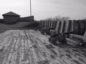 Historic cannons displayed along wooden fortification walls with a building visible in the background.