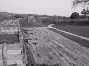 Aerial view of wooden fortification walls with mounted cannons overlooking a winding path.