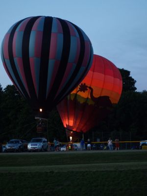 Hot air balloons illuminated at night on a field with spectators gathering around.