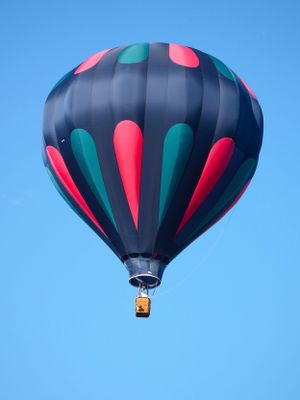 A black and red striped hot air balloon soars through the bright blue sky during a festival.