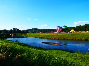 Hot air balloons float over a scenic landscape with a lake and rolling hills in the background.