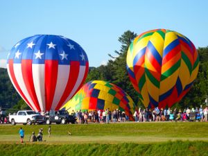 Row of inflated patriotic and multicolored hot air balloons on a grassy field with spectators.