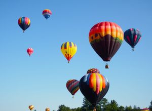 Multiple hot air balloons in flight showing various patterns and bright colors against blue sky.