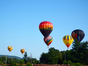 Multiple vibrant hot air balloons float against a bright blue sky over a mountain landscape.
