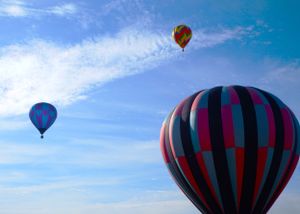 Three hot air balloons with bold stripes and patterns drift through wispy clouds.