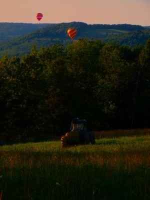Hot air balloons drift over a rural landscape with a tractor in the field at sunset.