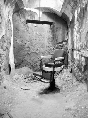 Abandoned prison cells with old barber chairs in a deteriorating concrete corridor.