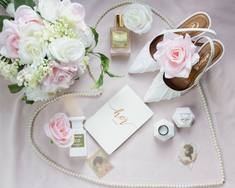 Elegant flatlay with pink and white roses, perfume bottle, pearls and beauty products arranged on soft pink fabric.