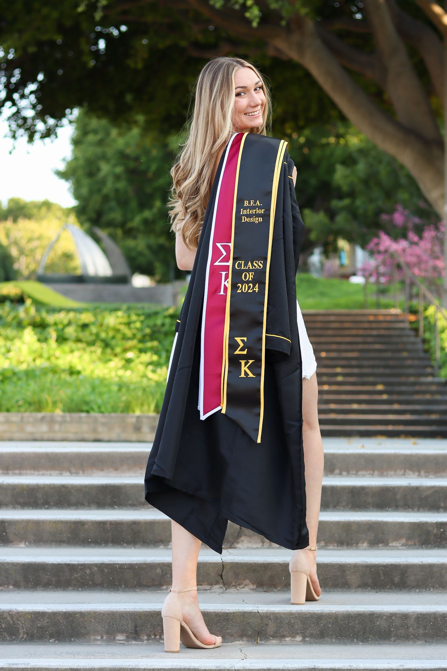 Graduate in black gown and honor cords poses on stone steps outdoors.