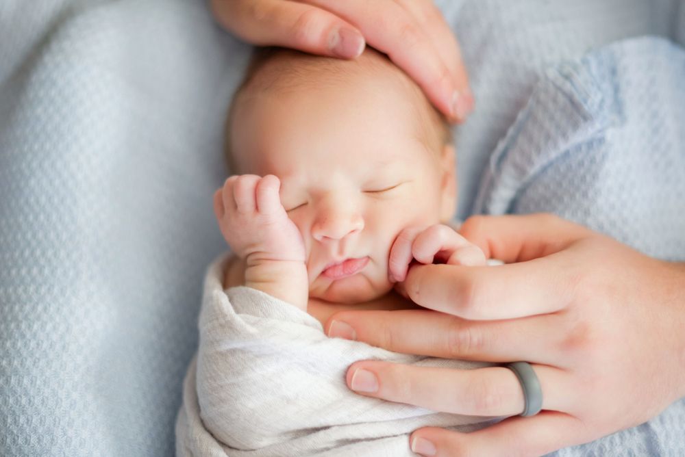 A sleeping newborn baby in white clothing is gently being held by caring hands against a soft blanket.