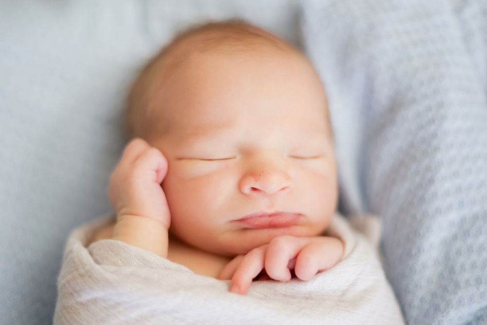 Sleeping newborn baby swaddled in soft grey blanket rests peacefully with tiny hands near face.