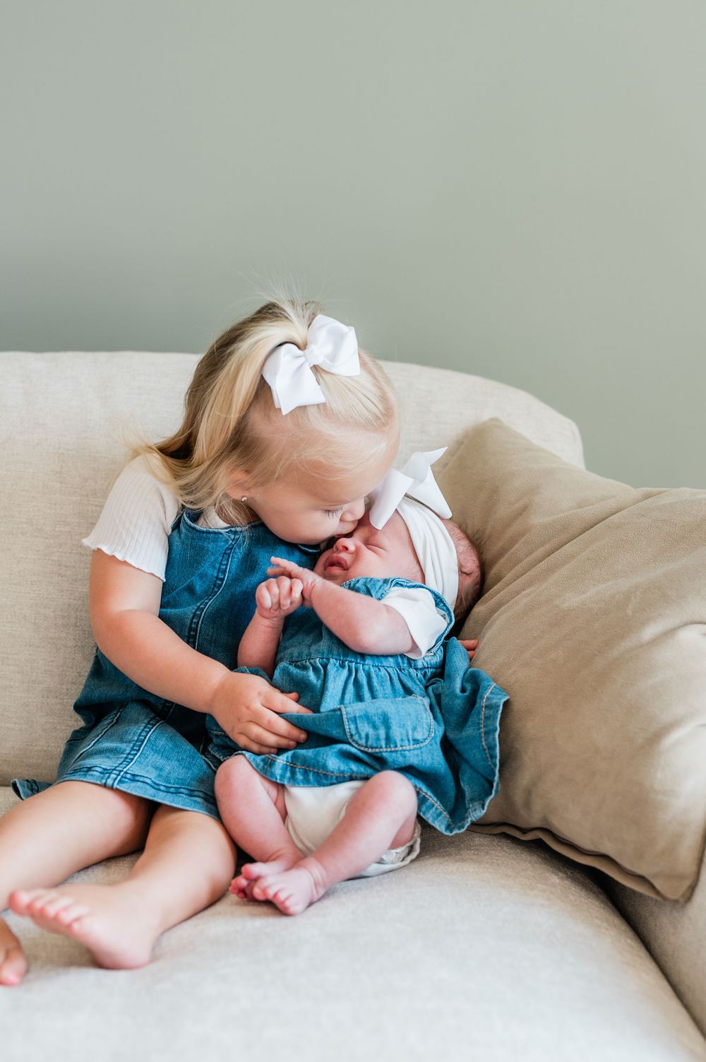 Toddler girl in denim dress with white bow kisses newborn baby sibling on a beige couch.