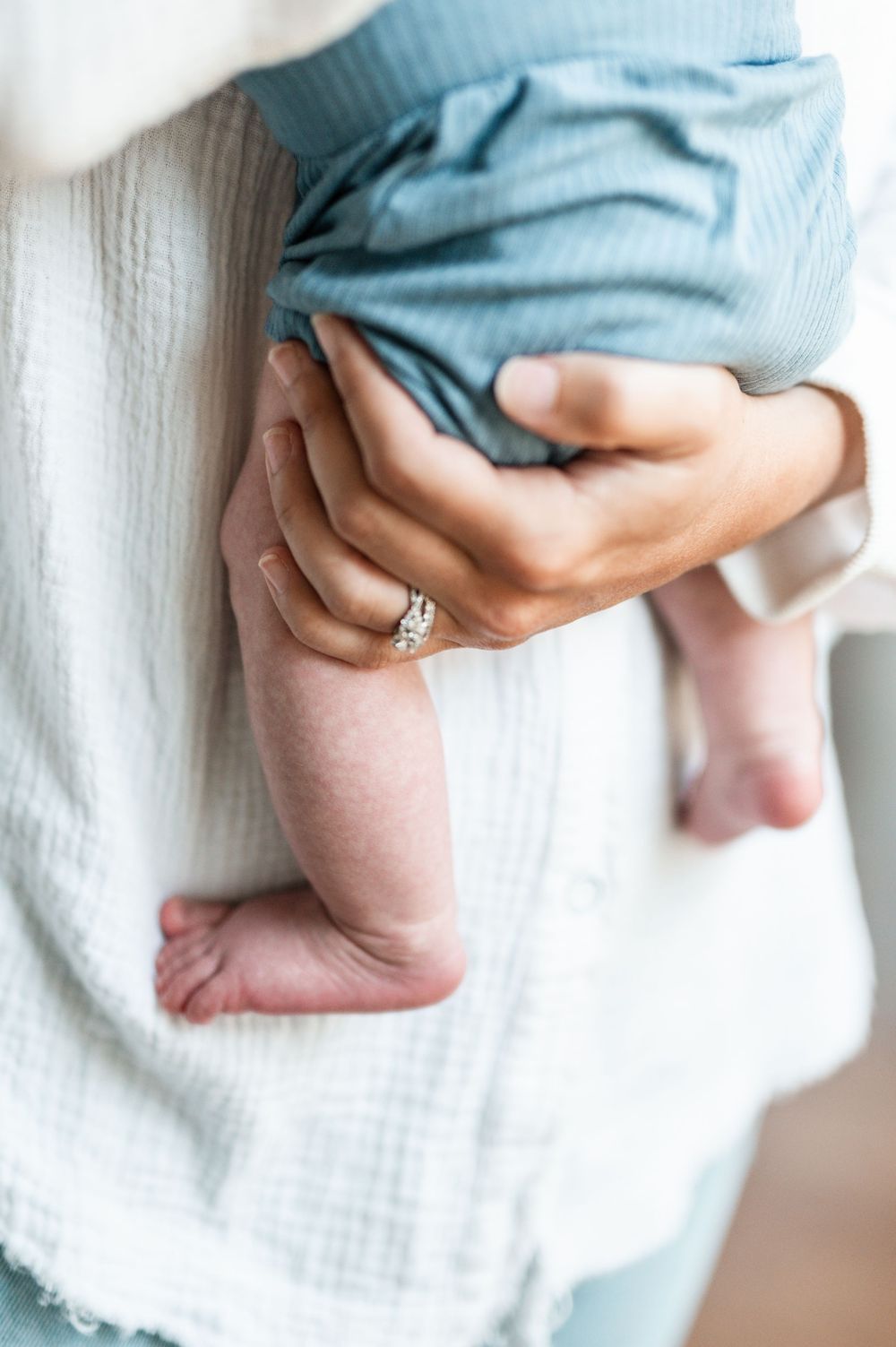 Close-up of a mother's hand with diamond ring cradling a newborn baby's legs wrapped in a soft white muslin blanket.