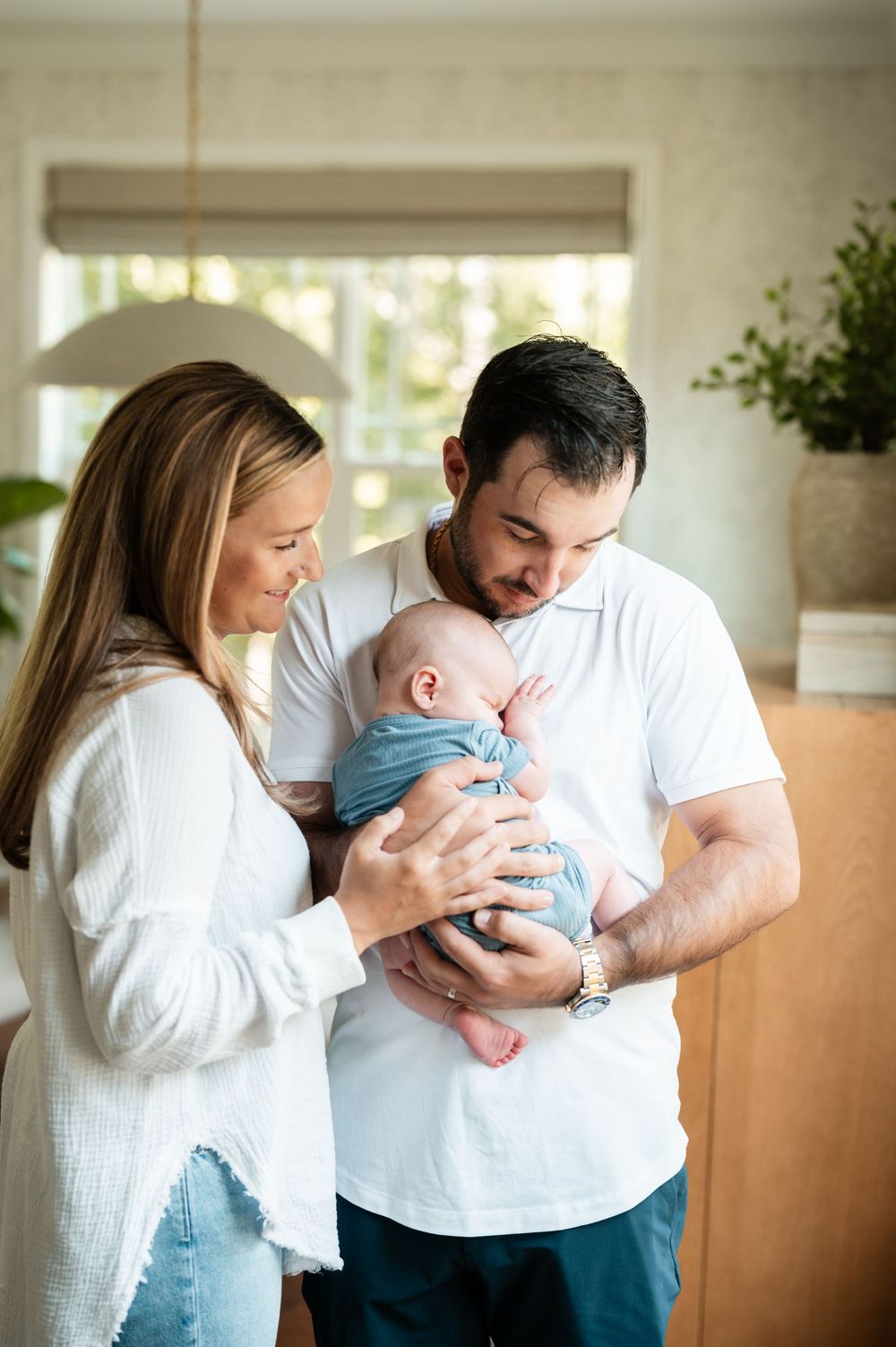 A couple in white tops lovingly gazes at their newborn baby wrapped in a blue swaddle indoors.