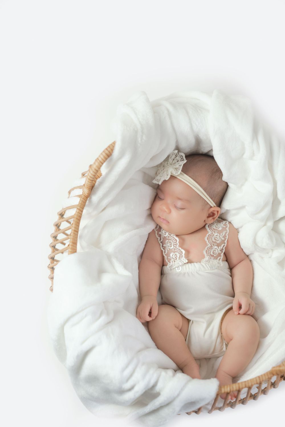 Peaceful sleeping newborn wearing white lace outfit lying on soft white blanket in woven basket.