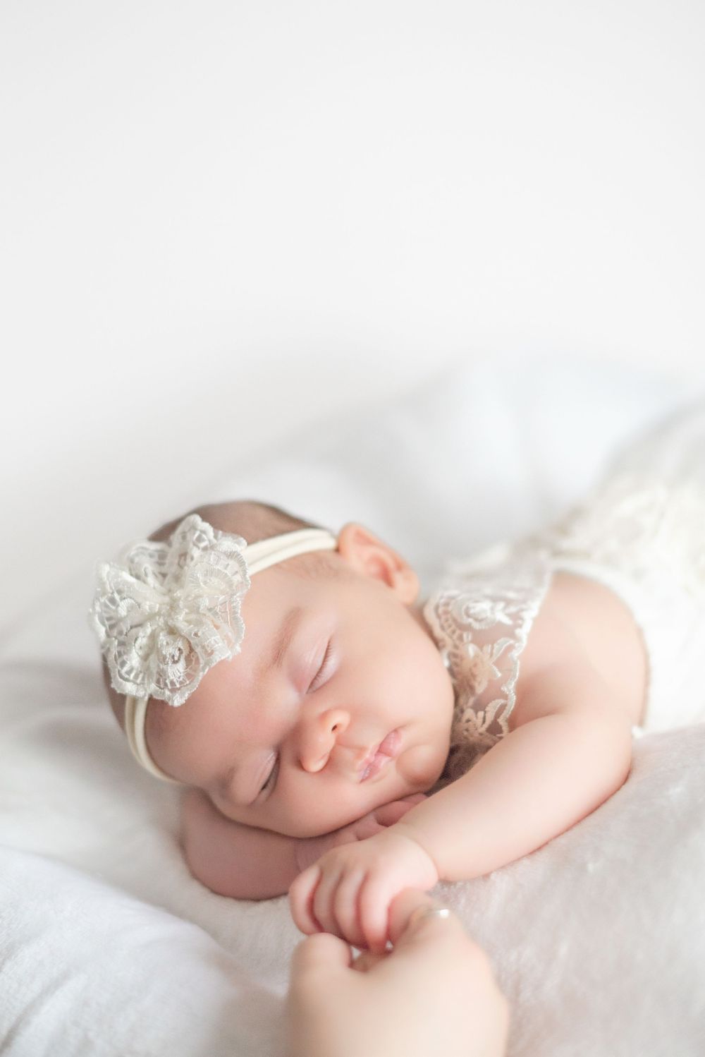 Sleeping baby wearing delicate white lace headband rests peacefully on soft white bedding.