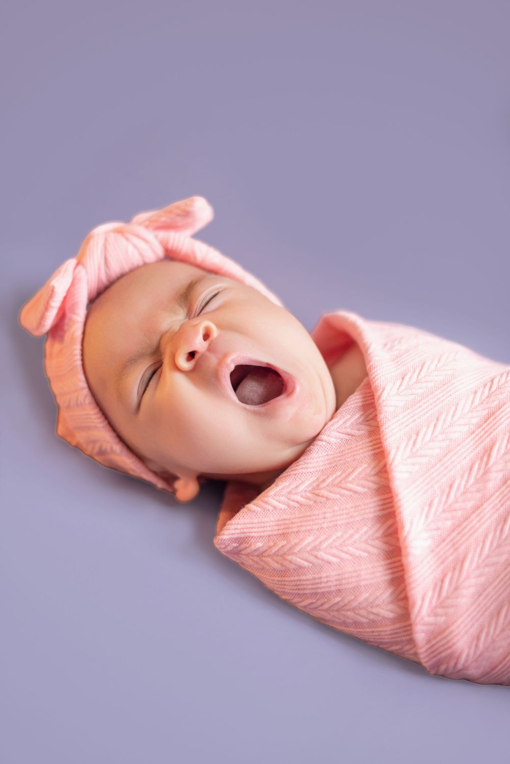 Yawning newborn swaddled in soft pink blanket with matching bow headband on purple background.
