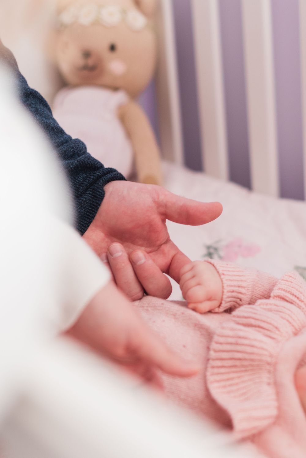 Close-up view of tiny baby hand touching soft pink knitted fabric in a crib with blurred plush toy in background.