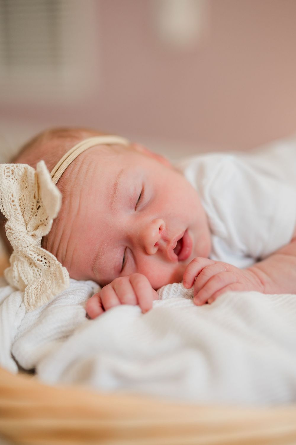 Sleeping newborn baby girl wearing a lace bow headband, wrapped in a white blanket in a cozy basket.