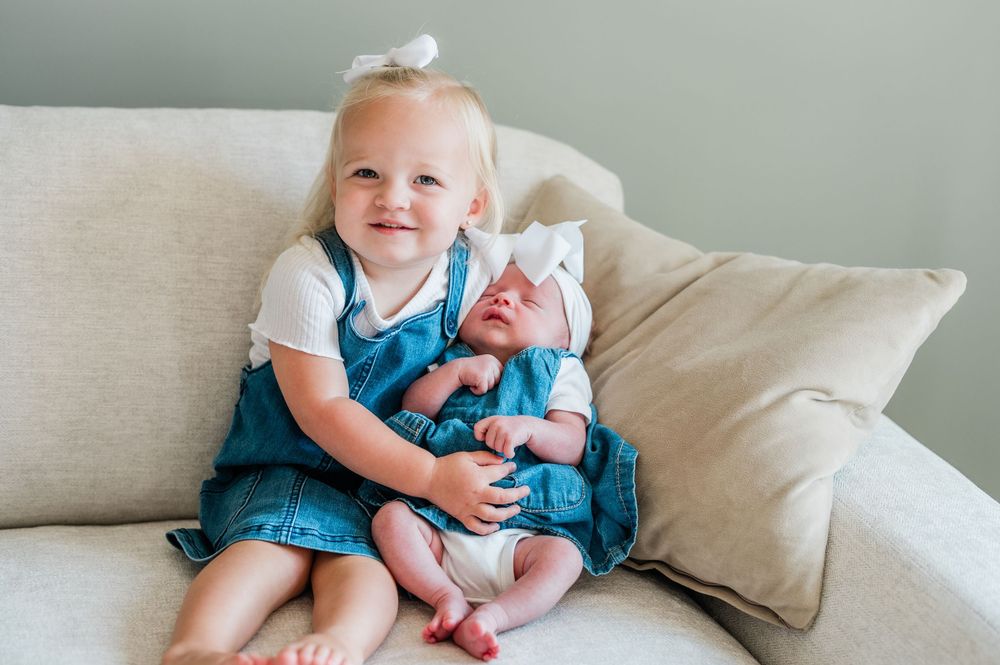Toddler girl in denim dress holding newborn baby sibling on a beige couch, both wearing white bows.