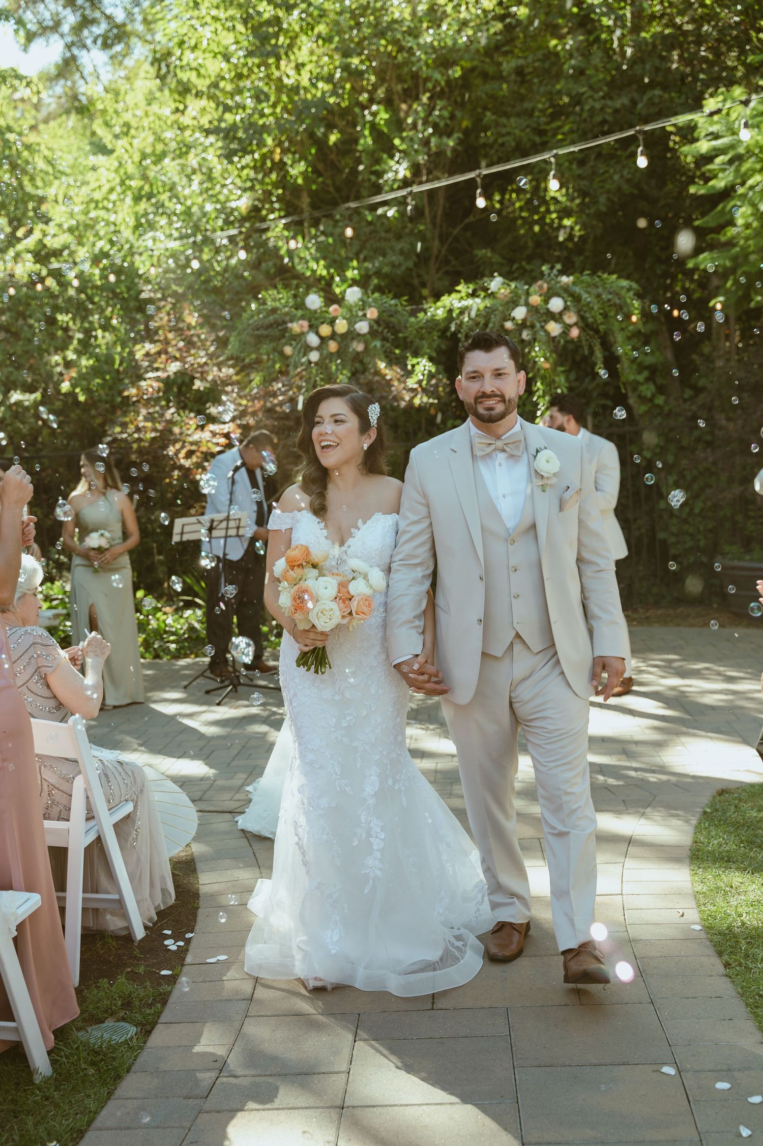 A joyous wedding exit as the newlyweds walk down an outdoor aisle under string lights and greenery.
