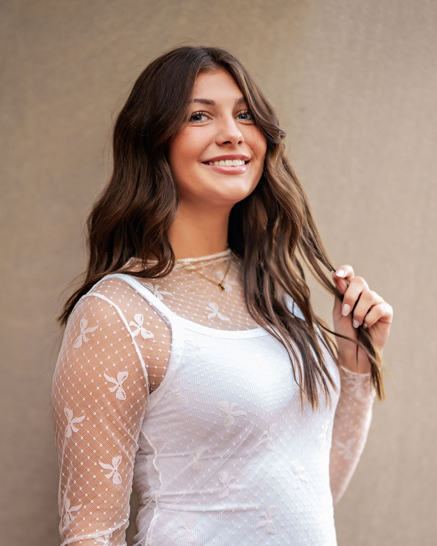 Young woman in a white lace long sleeve dress with sheer mesh detailing smiles while playing with her wavy brown hair.