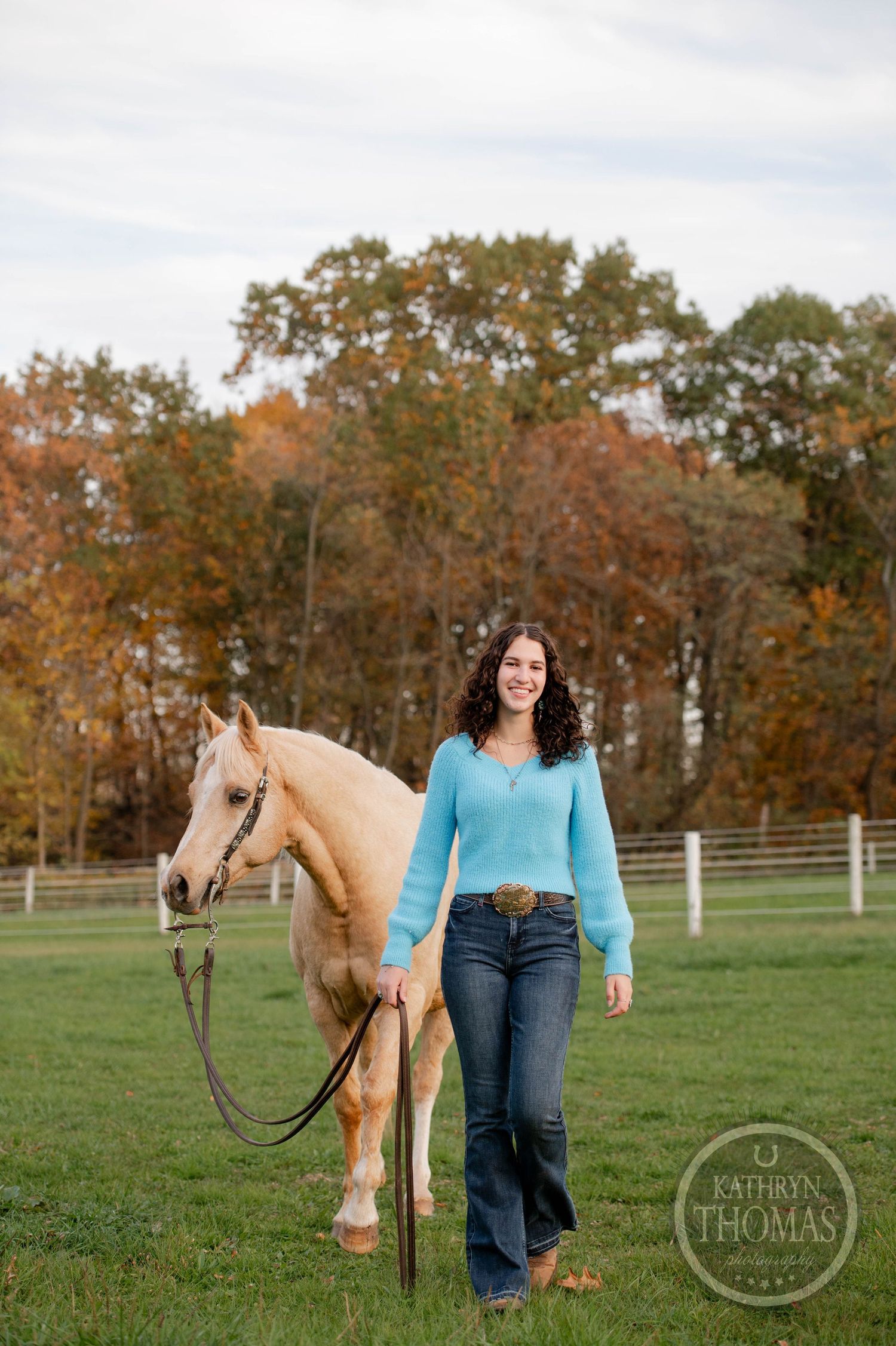A cream-colored horse with its handler stands in a fenced pasture during fall season.