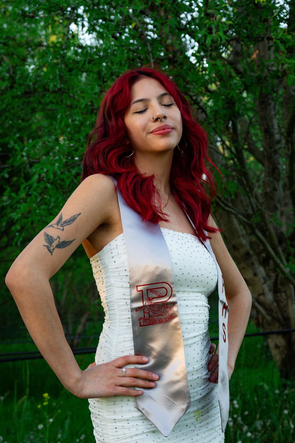Graduate in white dress and silver stole poses against green foliage background.