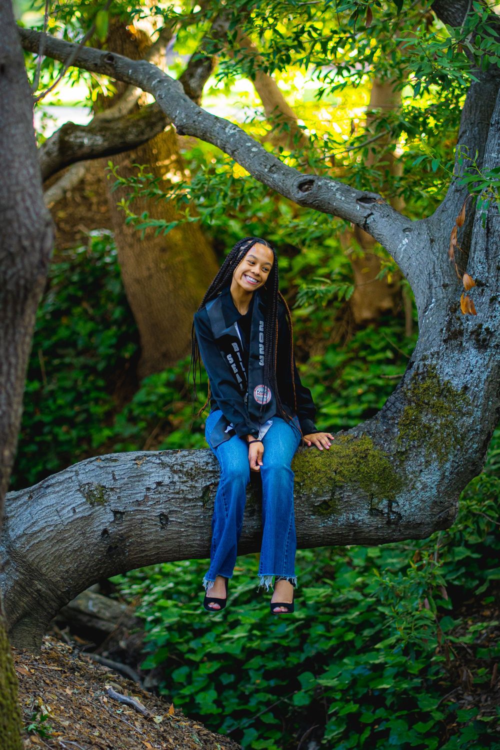 Graduate sitting on large tree branch in natural outdoor setting.