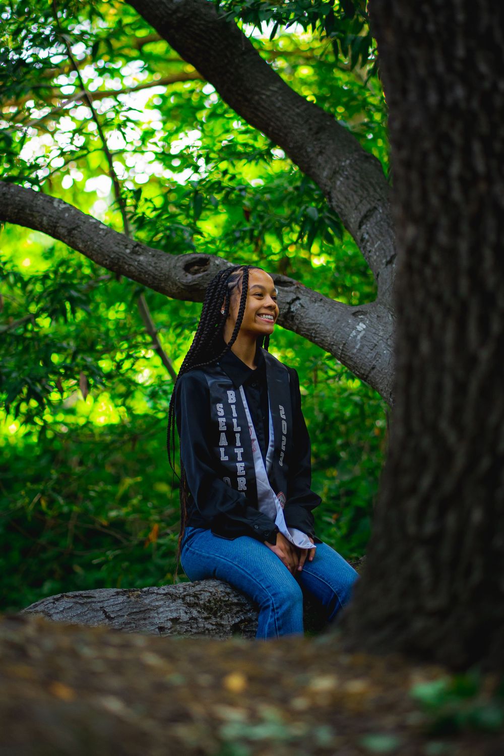 Graduate sitting on a tree branch in a wooded area wearing jeans and a dark hoodie.