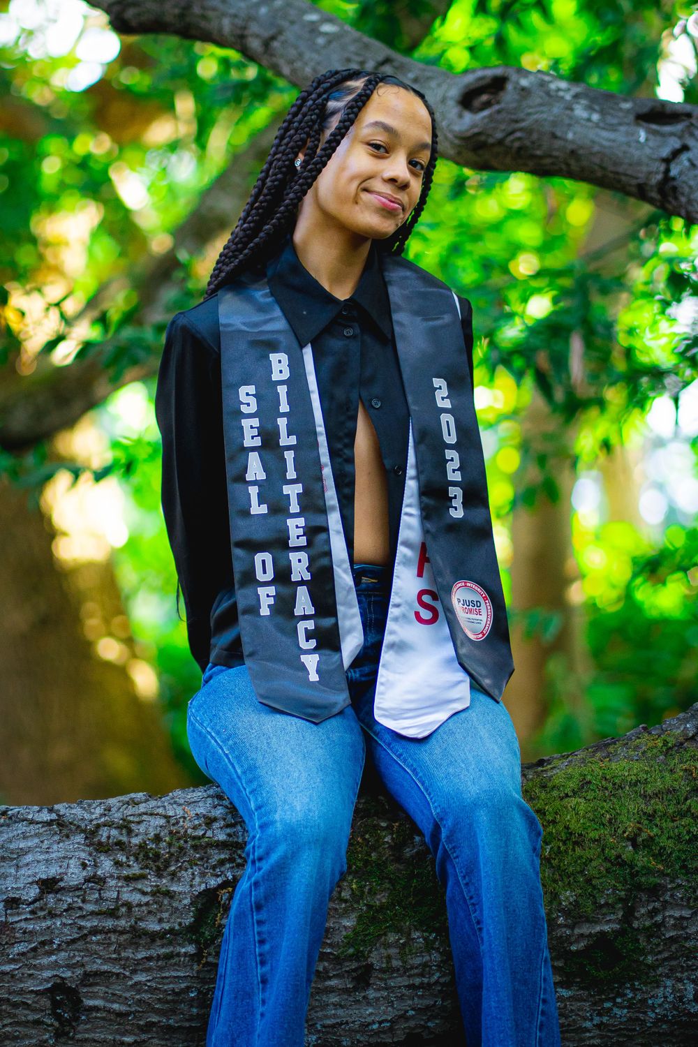 Graduate posing on tree trunk wearing graduation stoles and casual attire outdoors.