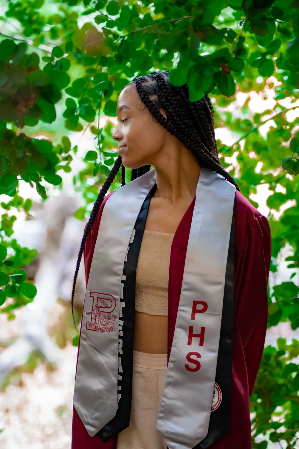 Graduate wearing maroon and silver graduation stoles smiling against lush green foliage.
