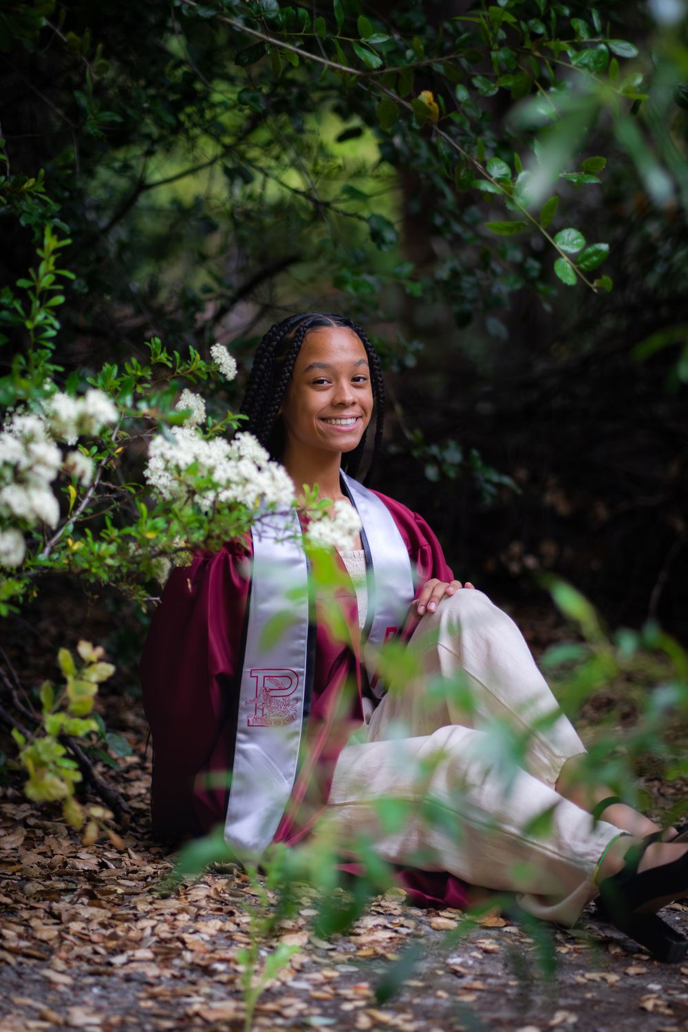 Graduate in maroon regalia seated among blooming white flowers in a garden setting.