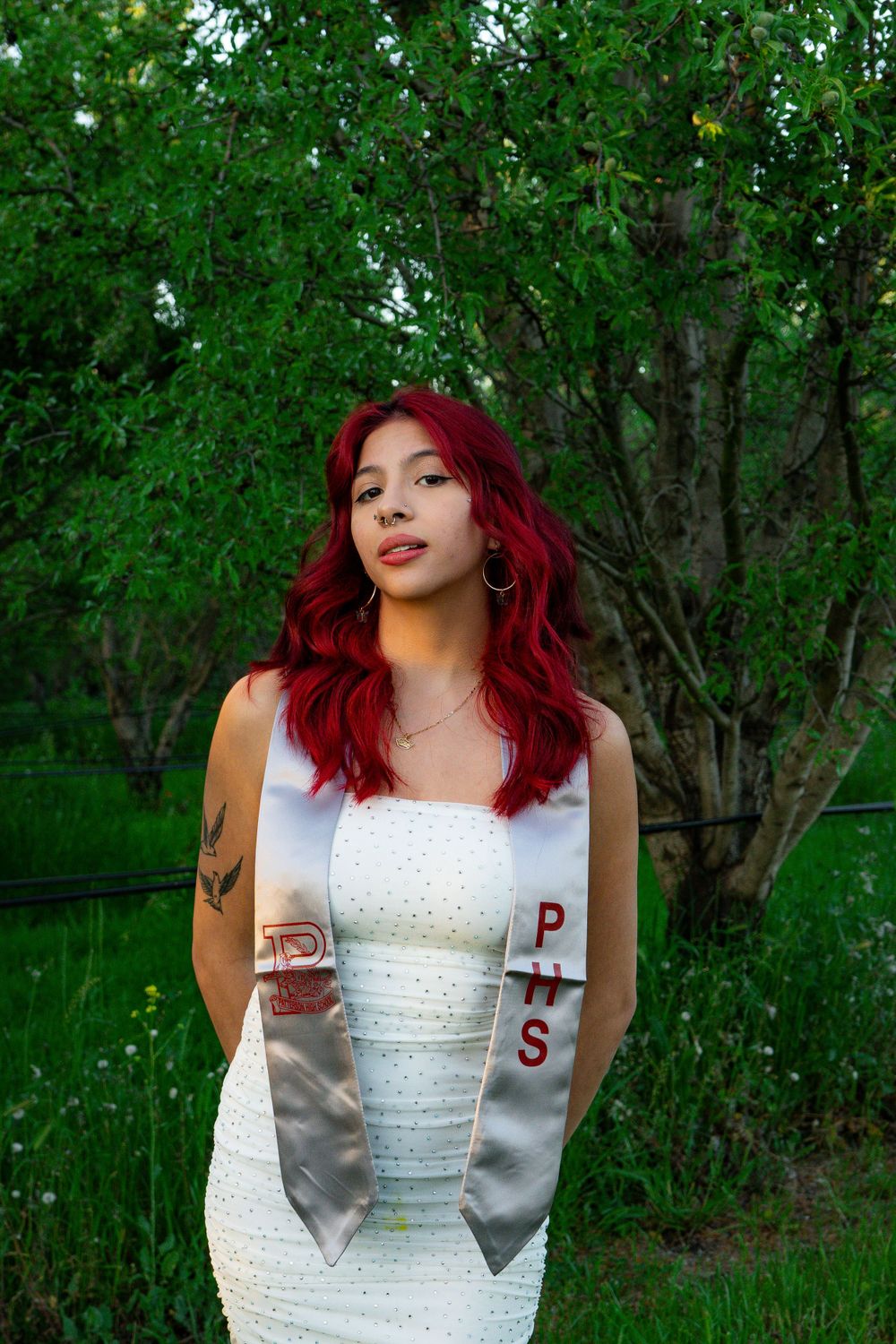Graduate in elegant white dress and academic stoles standing in lush garden setting.