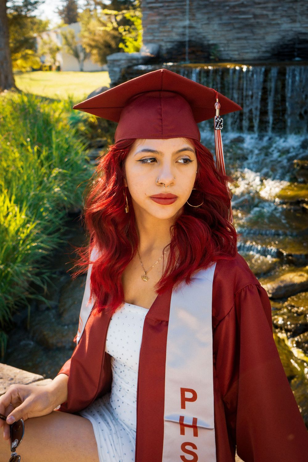 Graduate in maroon cap and gown poses by waterfall in sunlit outdoor setting.