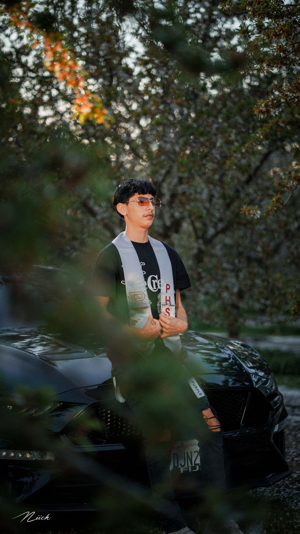 Portrait through tree branches beside black car wearing casual streetwear.