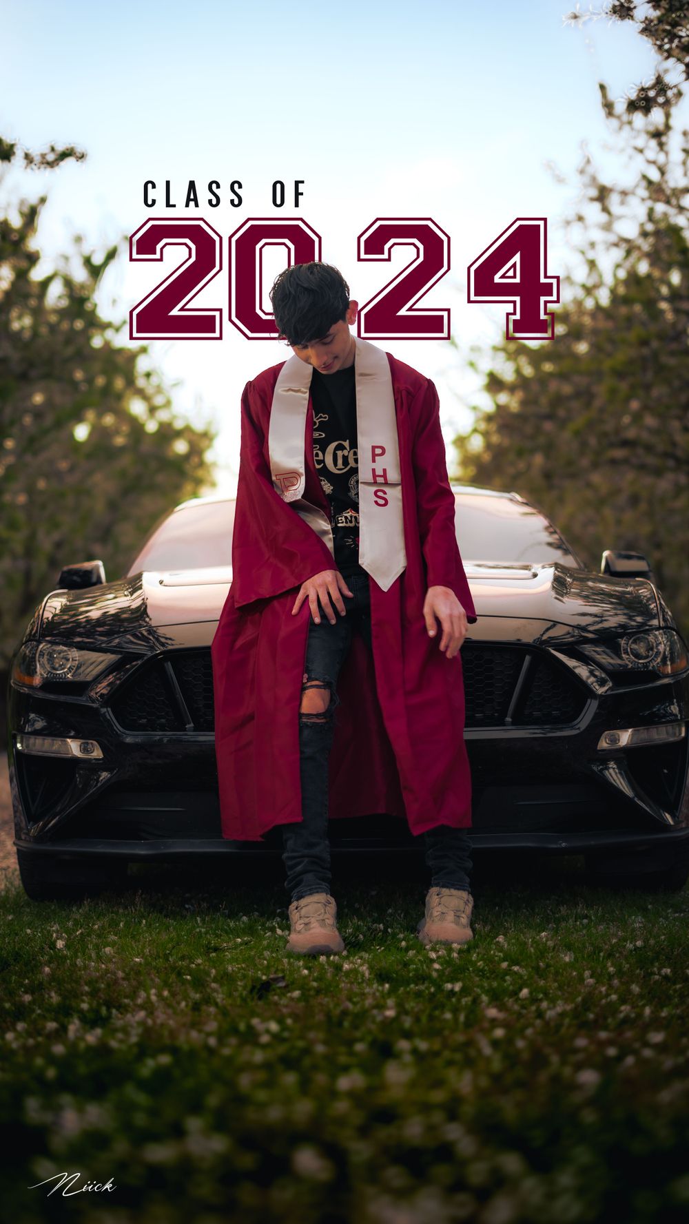 Class of 2024 graduate in red gown posing on black Mustang car.