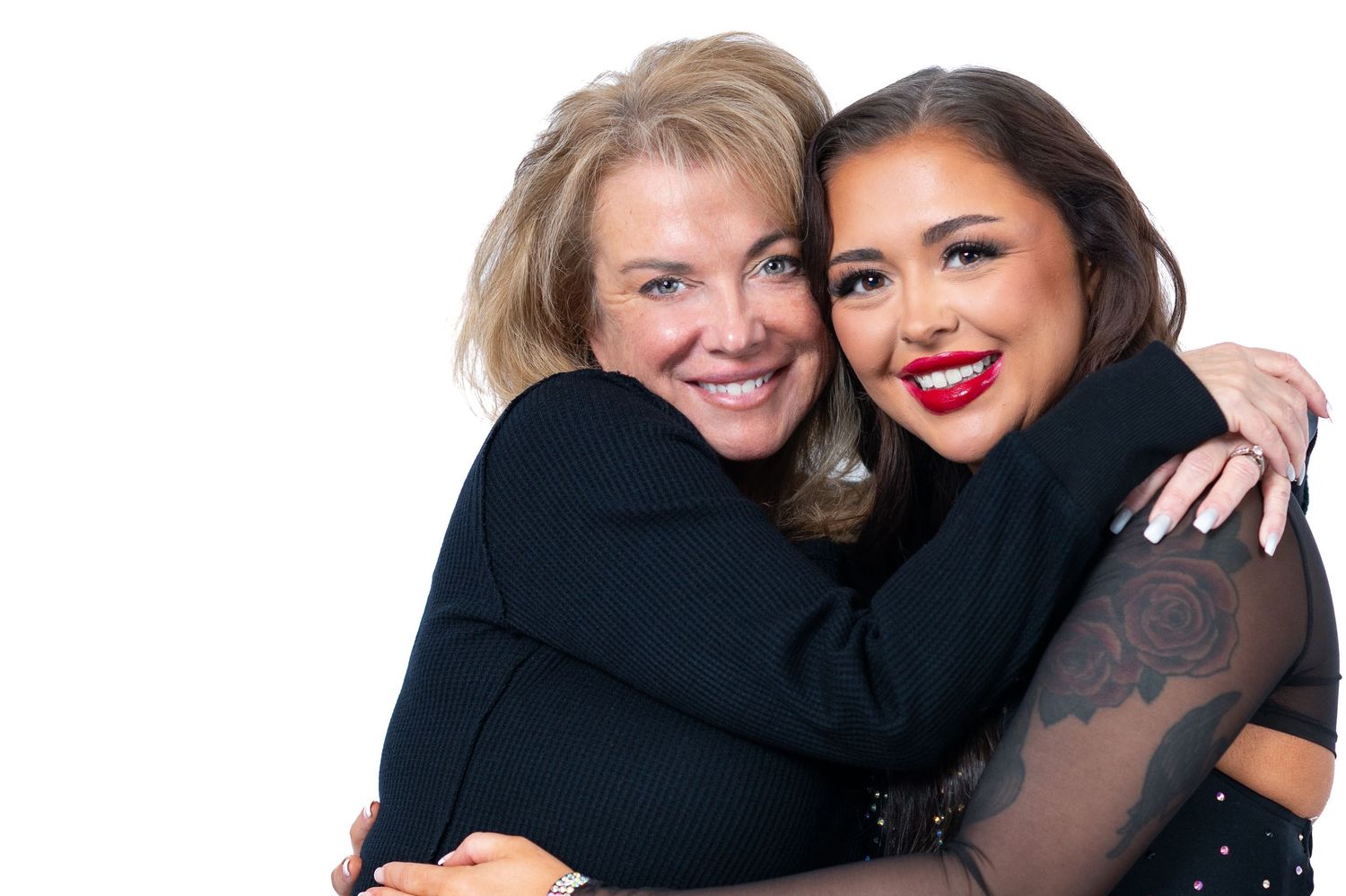 Two women in black tops embrace and smile warmly against a white background.