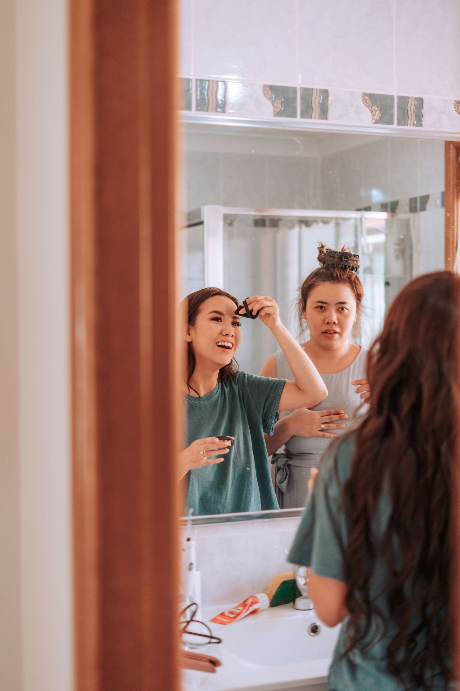 Two people applying makeup and getting ready in front of a bathroom mirror with a joyful atmosphere.