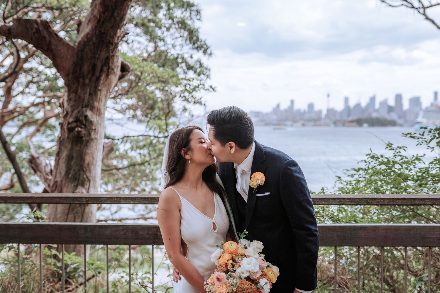 Romantic wedding couple embracing on waterfront balcony with city skyline and harbor view in background.